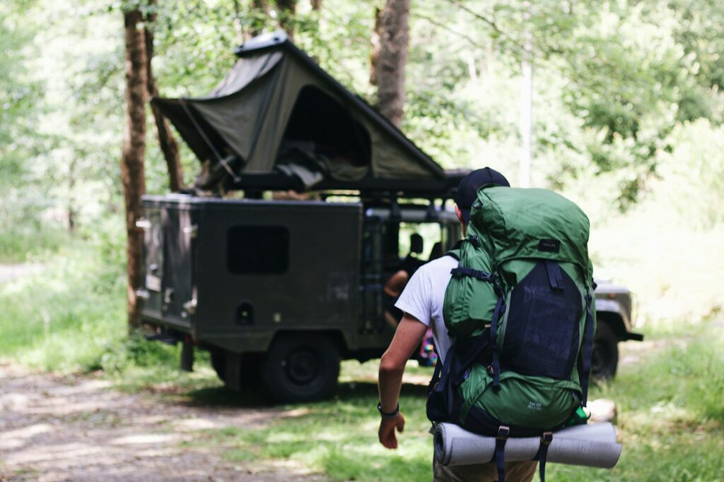 A person with a large green backpack walks towards a rugged vehicle with a tent, surrounded by a lush forest.