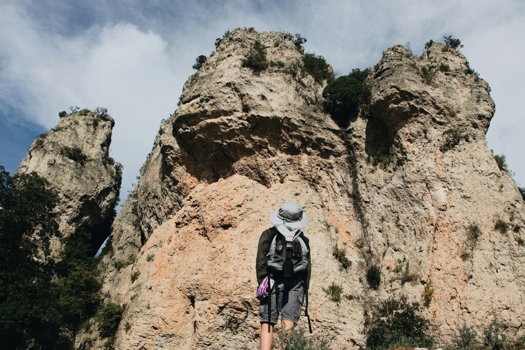 A hiker stands before towering rocky cliffs, gazing up at the natural formations under a partly cloudy sky.