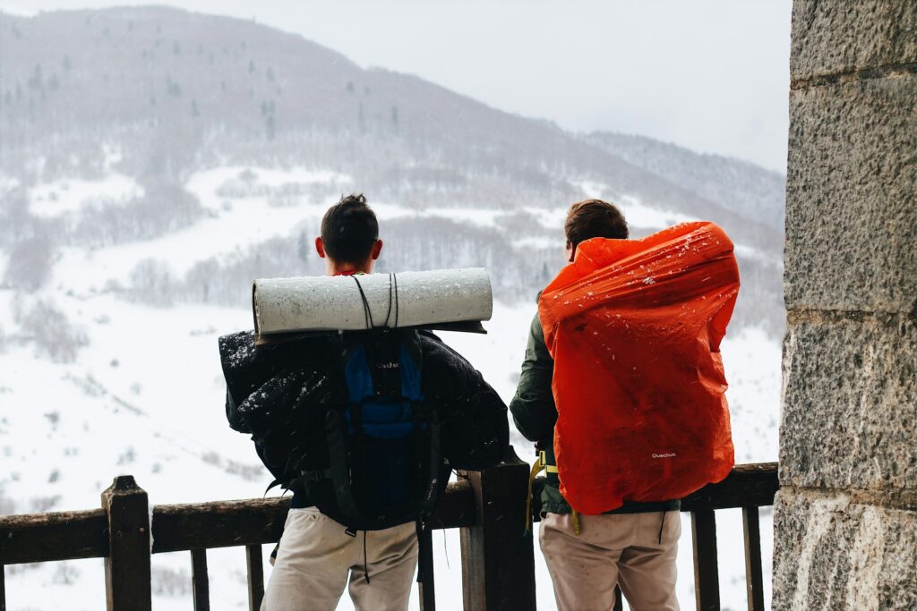 Two hikers with backpacks and sleeping mats stand on a snowy overlook, gazing at distant mountains under a cloudy sky.