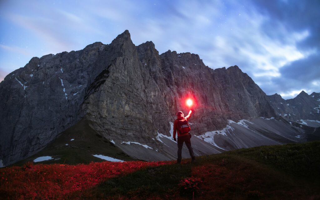 A lone hiker in a red jacket signals with a red flare against a dramatic mountain backdrop under a twilight sky.