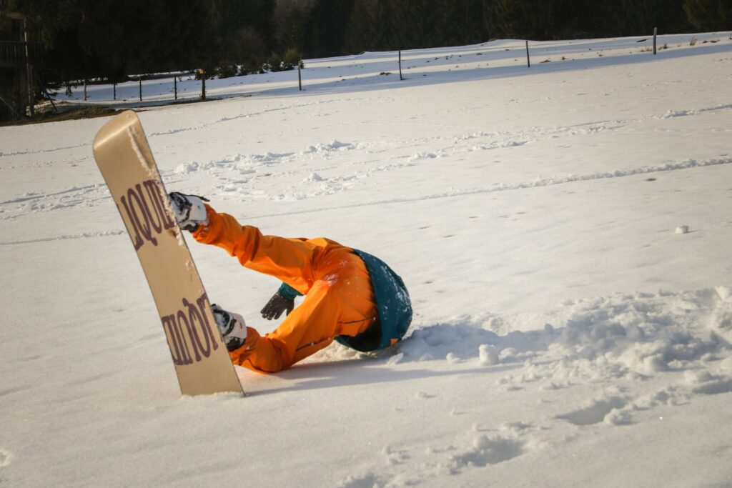 A snowboarder in an orange outfit tumbles face-first into a snowy field, with snow scattered around and trees in the background.