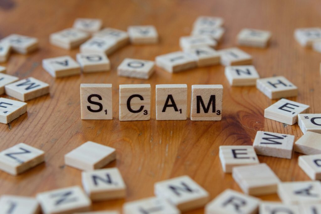 Four wooden Scrabble tiles spell "SCAM" on a wooden table, surrounded by scattered tiles with letters.