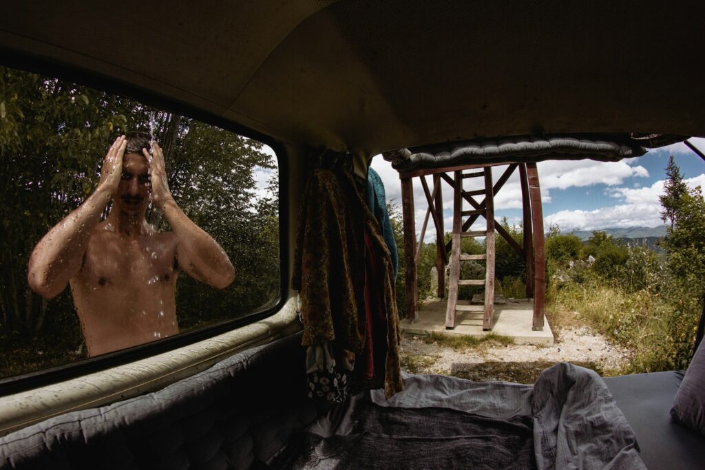 A view from inside a van showing a rain-splattered window, outdoor scenery, and a wooden structure in the distance.
