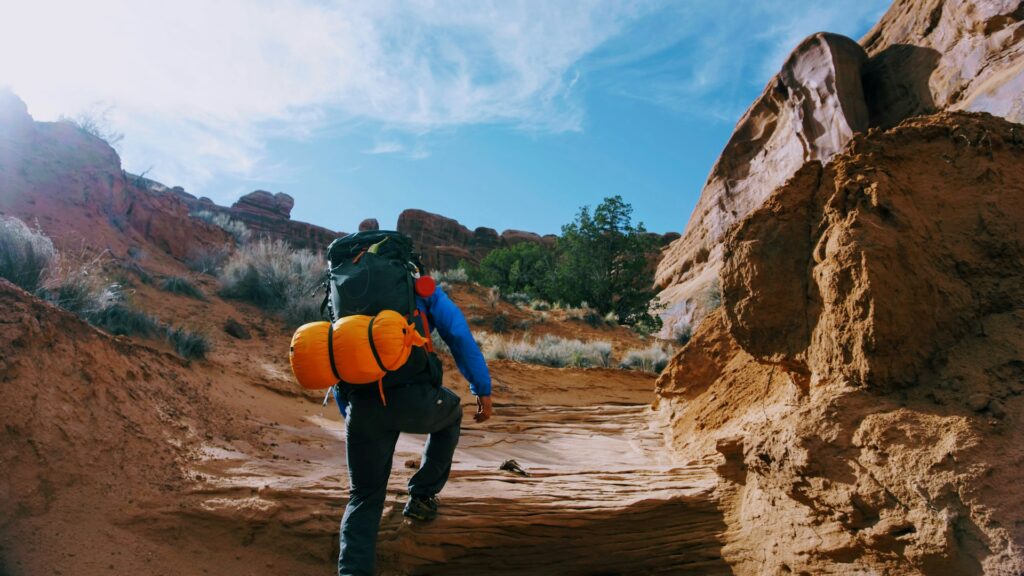 A hiker climbs a rocky trail, carrying a large backpack with a bright orange sleeping bag, under a blue sky with wispy clouds.