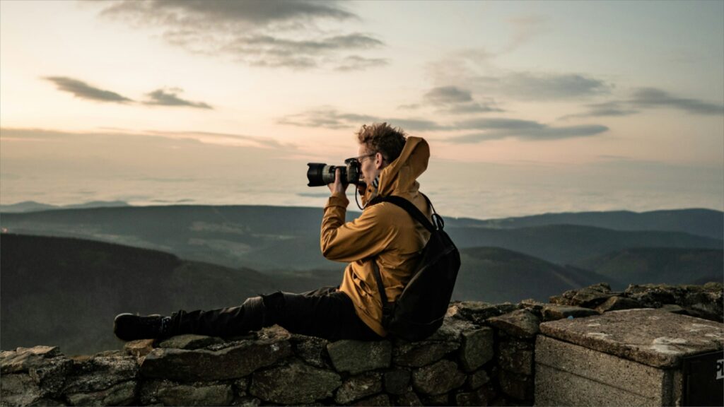 A man sits on a rock, capturing a photo of the majestic mountains in the background.