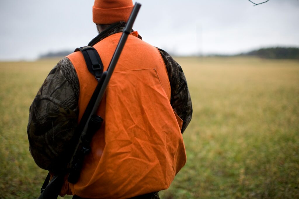 A hunter in an orange vest and hat stands in a field, holding a rifle, preparing for a hunting trip on a cloudy day.