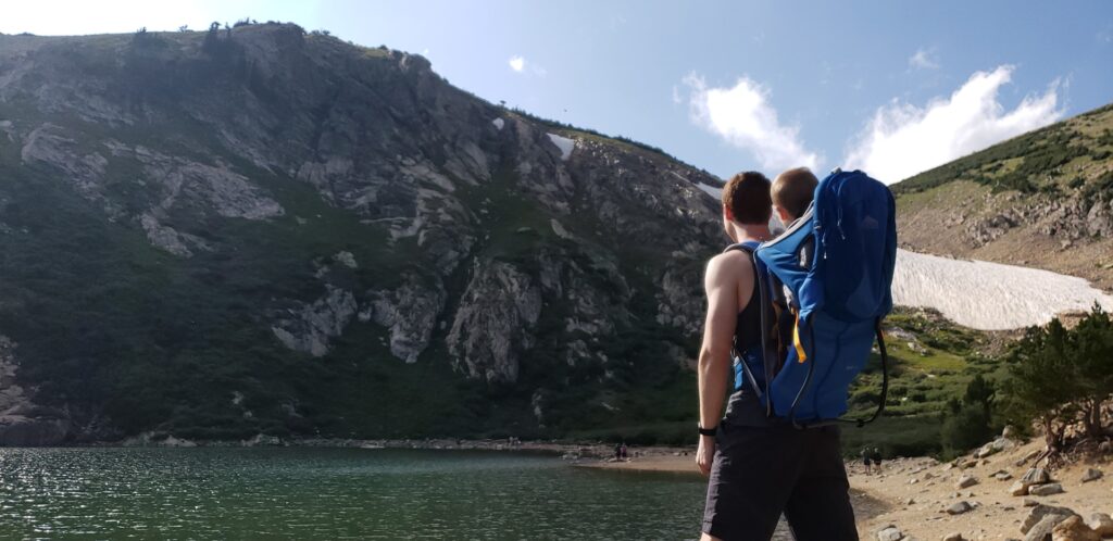 A hiker with a child in a backpack gazes at a towering mountain, near a serene lake surrounded by lush greenery and snow patches.