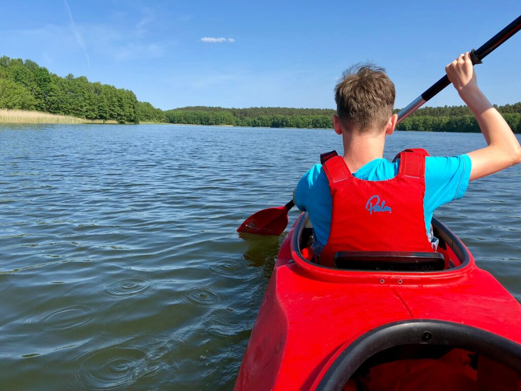 A person in a red kayak paddles on a serene lake surrounded by lush greenery under a clear blue sky.