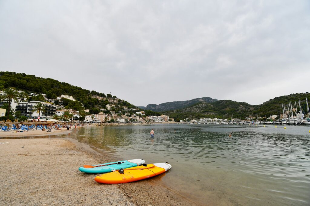 Two paddleboards in turquoise and orange on a tranquil beach with light crowds, surrounded by hills and coastal buildings.