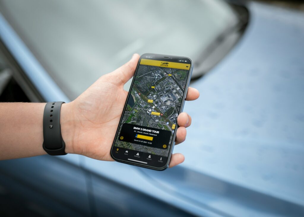 A person holds a smartphone displaying a map for the "BMW 2 Grand Tour" event, with a car visible in the background.
