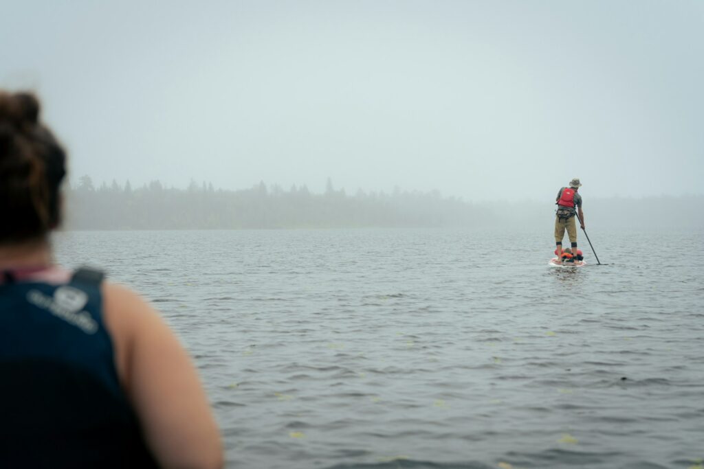 A person paddleboarding in a foggy lake, viewed from behind a seated individual, with trees faintly visible in the misty background.