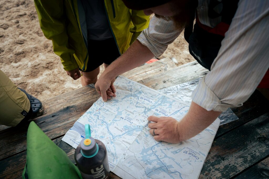 Two individuals study a detailed map on a wooden table, surrounded by sandy terrain, preparing for an outdoor adventure.