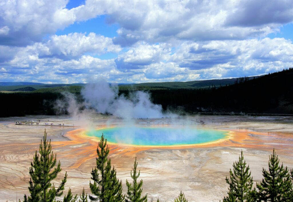 Aerial view of Yellowstone's Grand Prismatic Spring, showcasing vibrant blue and orange hues amidst steam and lush greenery under a cloudy sky.