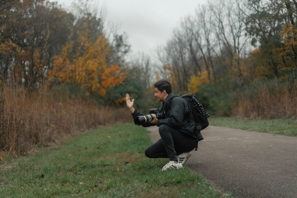 A photographer crouches on a path, holding a camera while surrounded by autumn foliage and overcast skies.