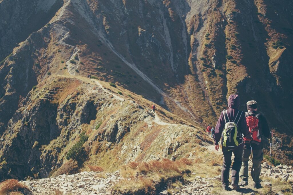 Two hikers, one in a green backpack and one in a red, walk along a rocky mountain path with others in the distance under clear skies.