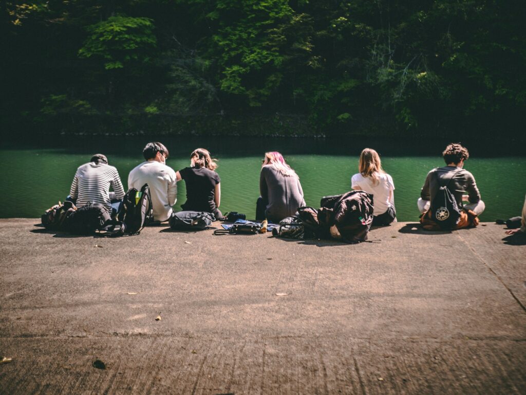 A group of six people sitting by a calm green river, backpacks beside them, enjoying the peaceful outdoor scenery.