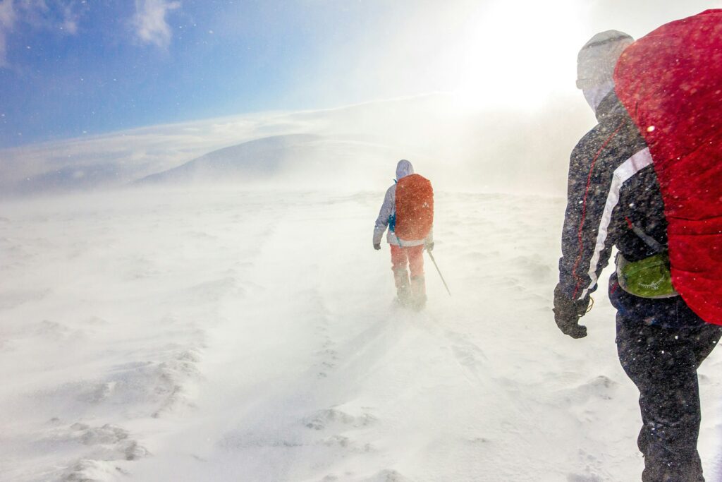 Two hikers traverse a snowy landscape, battling strong winds and limited visibility under a bright, blinding sun.