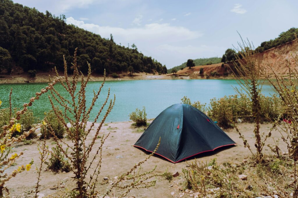 A black camping tent beside a serene turquoise lake, surrounded by green hills and wild plants under a partly cloudy sky.