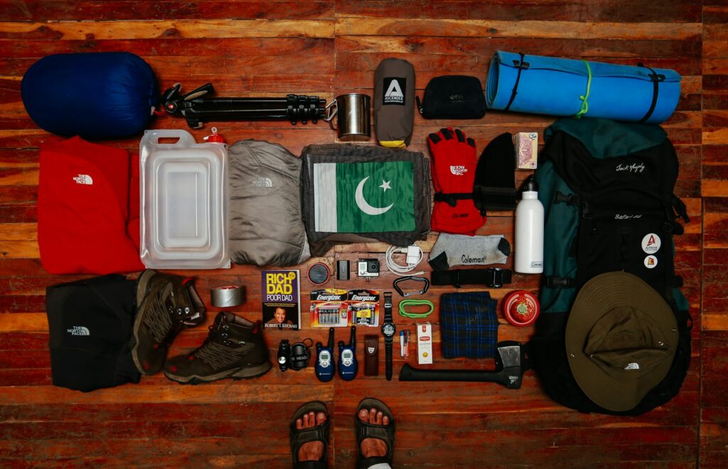 A flat lay of camping gear includes a tent, sleeping bag, cooking utensils, a backpack, and various tools and supplies, set on wooden flooring.