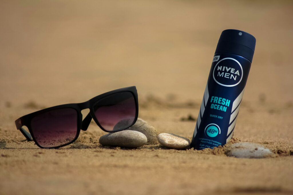 A black pair of sunglasses rests on sand alongside a Nivea Men Fresh Ocean deodorant, surrounded by smooth pebbles.