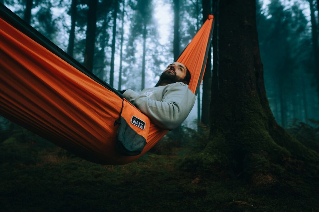 A person relaxes in an orange hammock suspended between trees in a misty forest, surrounded by lush greenery and soft lighting.