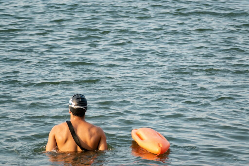 A man wearing a black swim cap is swimming in calm water, with an orange flotation device nearby.