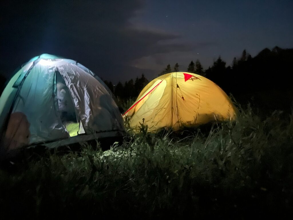 Two tents illuminated at night, one blue with a glowing interior and one yellow with a red marking, set in a grassy field.
