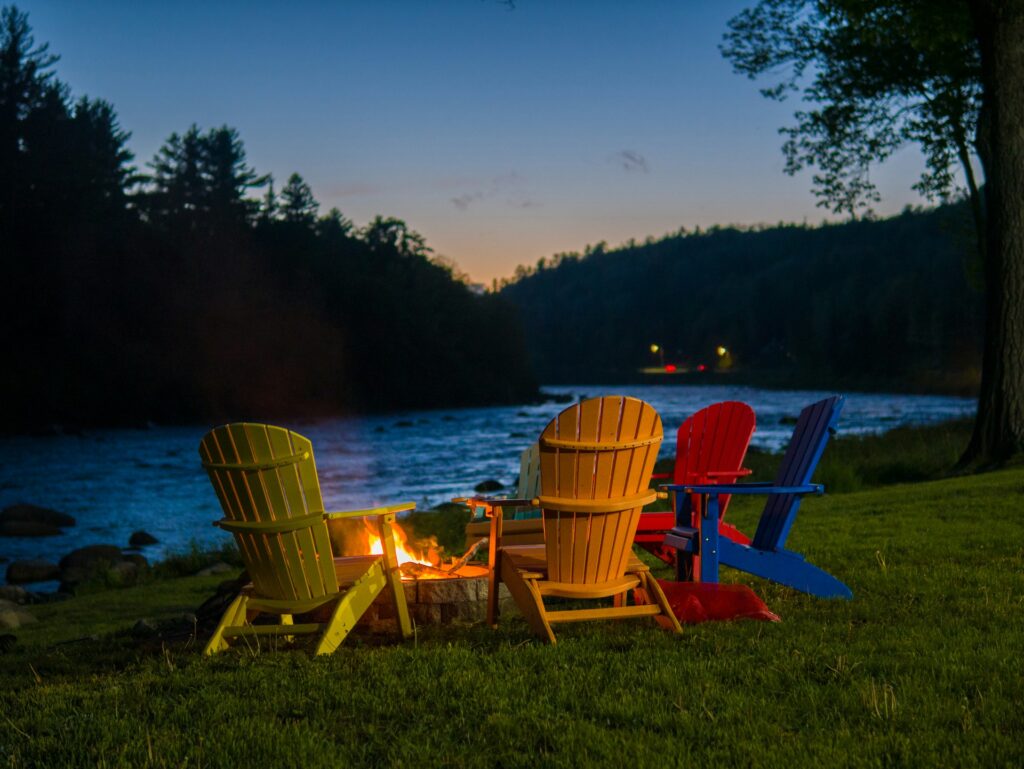 Colorful Adirondack chairs surround a fire pit by a river at dusk, with a serene forest backdrop and gentle water ripples.