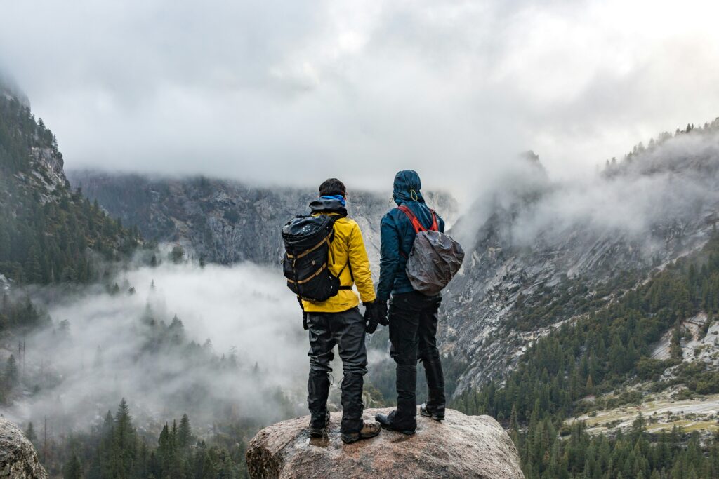 Two hikers in waterproof jackets stand on a rock, overlooking a misty forested valley surrounded by mountains.