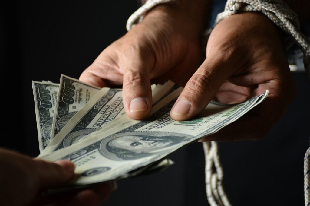 Two hands exchanging a stack of dollar bills, with one hand tied in rope, symbolizing a transaction under duress.