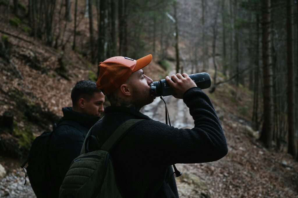 Two individuals in a forest, observing the surroundings while the other stands nearby, both wearing jackets.