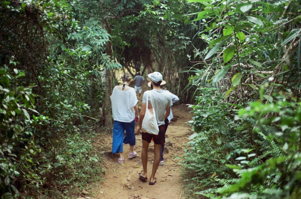 A group of people walking along a narrow trail surrounded by dense green foliage in a serene forest setting.
