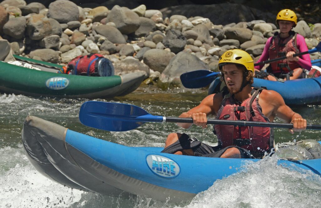 Two individuals in inflatable kayaks navigate a rocky river, paddling through splashing water on a sunny day.