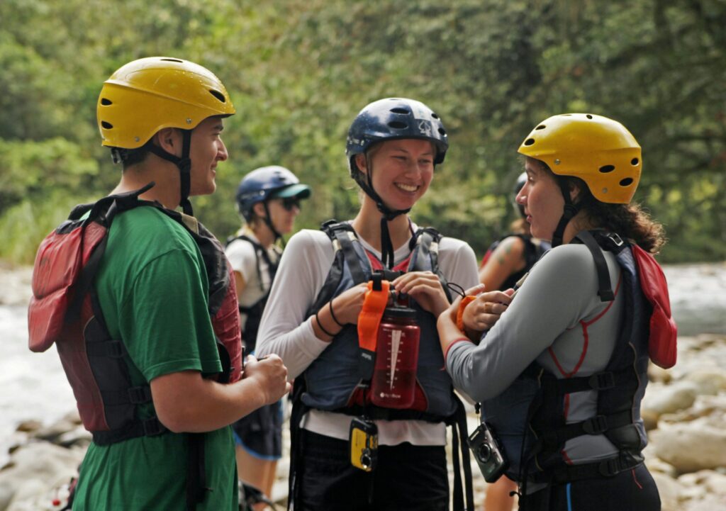 A group of people in helmets and life jackets gather near a river, discussing their rafting adventure amidst lush greenery.