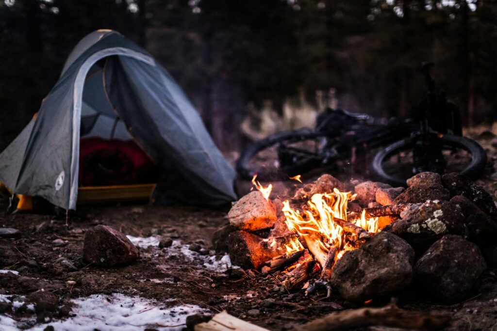 A camping scene featuring a blue tent, a glowing campfire with rocks, and a bicycle in the background amidst a forest setting.