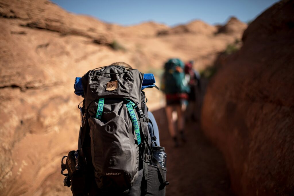 A hiker with a black backpack and blue sleeping bag walks through a narrow, rocky trail under a clear sky, with others in the background.