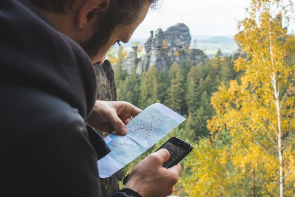A person examines a map while holding a smartphone, with a picturesque landscape of trees and rock formations in the background.