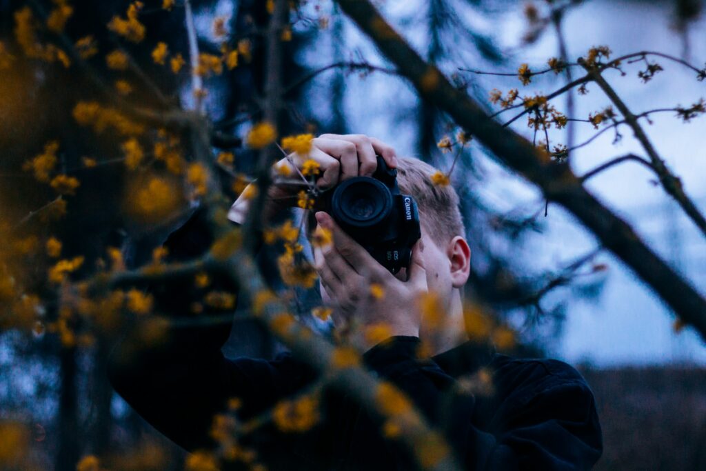 A young man with short hair holds a Canon camera, framed by branches and yellow flowers, capturing a moment outdoors.