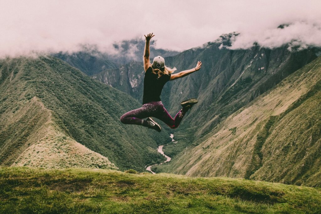 A person jumps joyfully on a mountain ridge, overlooking lush green valleys and hills under a cloudy sky.