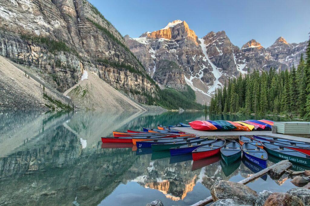 Colorful canoes line the shore of Moraine Lake, reflecting surrounding mountains and evergreen trees in the tranquil water.