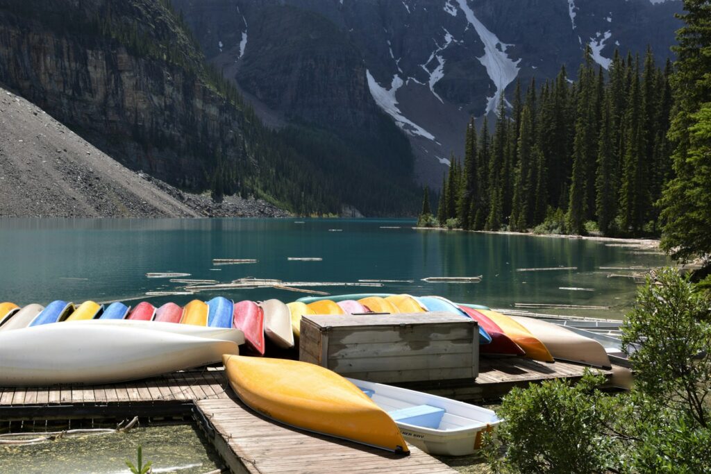 A vibrant assortment of kayaks rests on a wooden dock by a tranquil lake, surrounded by towering trees and majestic mountains.