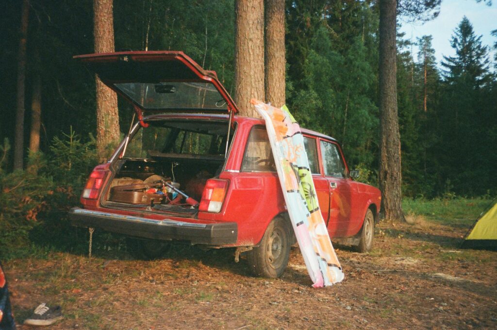 A red car parked in a forest, its trunk open with camping gear inside and a colorful towel leaning against it.