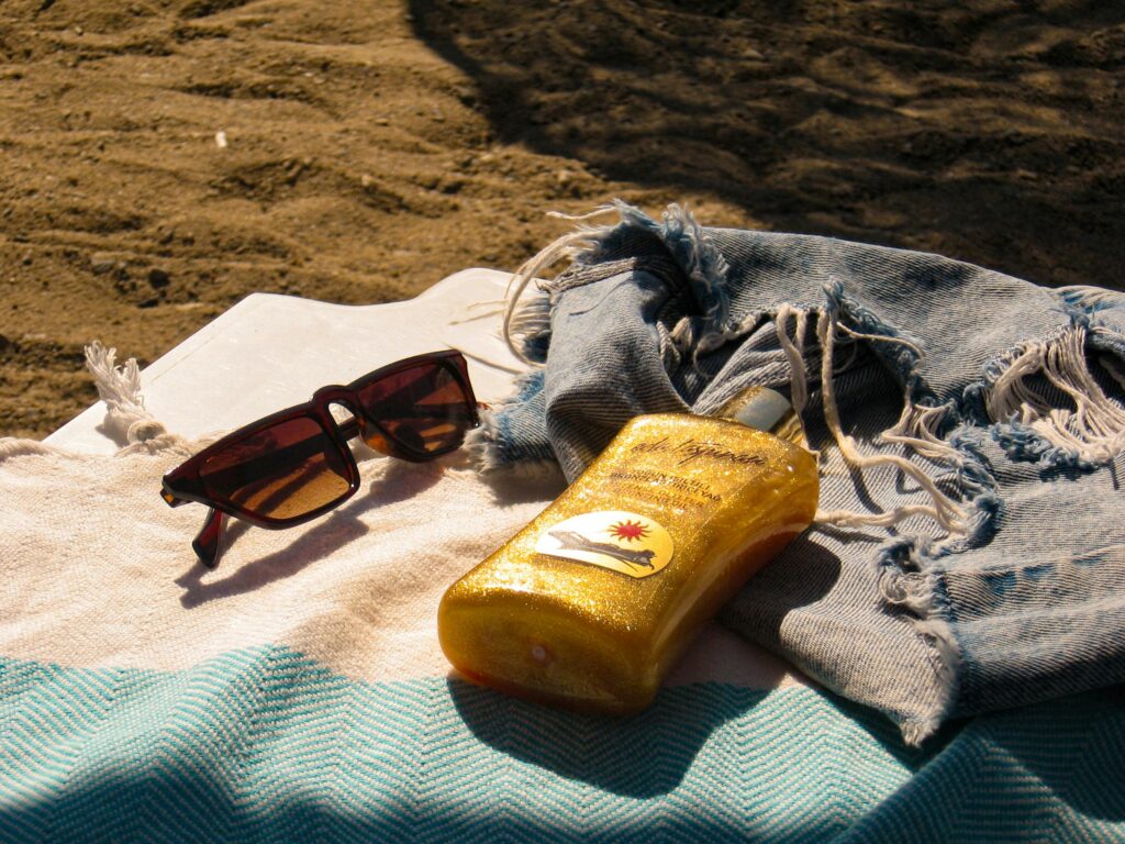 A pair of sunglasses rests on a textured towel alongside a bottle of golden sunscreen and a frayed denim jacket on the sandy beach.