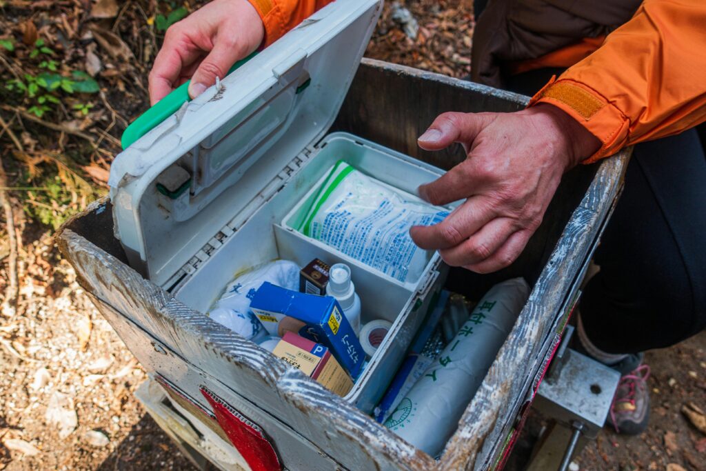 A person opens a first aid kit revealing various medical supplies, including ointments and bandages, while in a forested area.