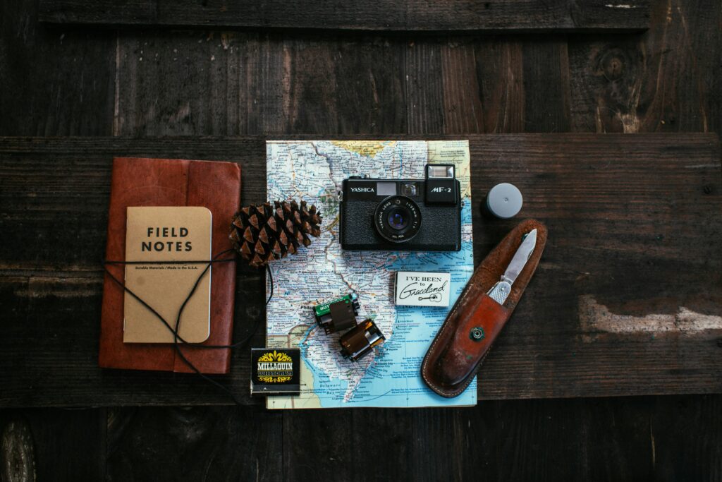 A vintage Yashica camera, field notes notebook, pinecone, and a folding knife are arranged on a map atop a wooden table.