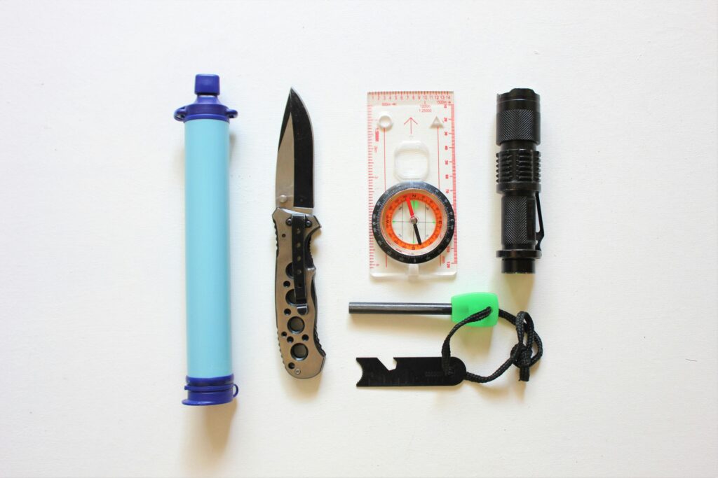A survival kit display featuring a water filter, folding knife, compass, flashlight, fire starter, and multi-tool on a white background.