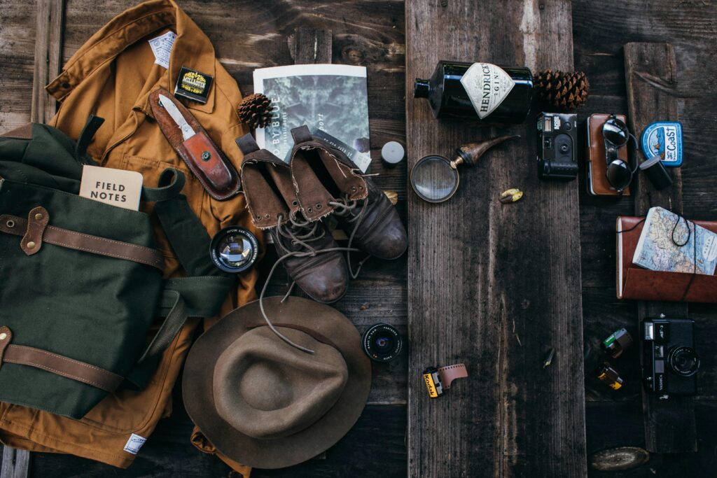 An arrangement of outdoor gear, including a jacket, boots, camera, flask, notebook, and map, on a rustic wooden surface.