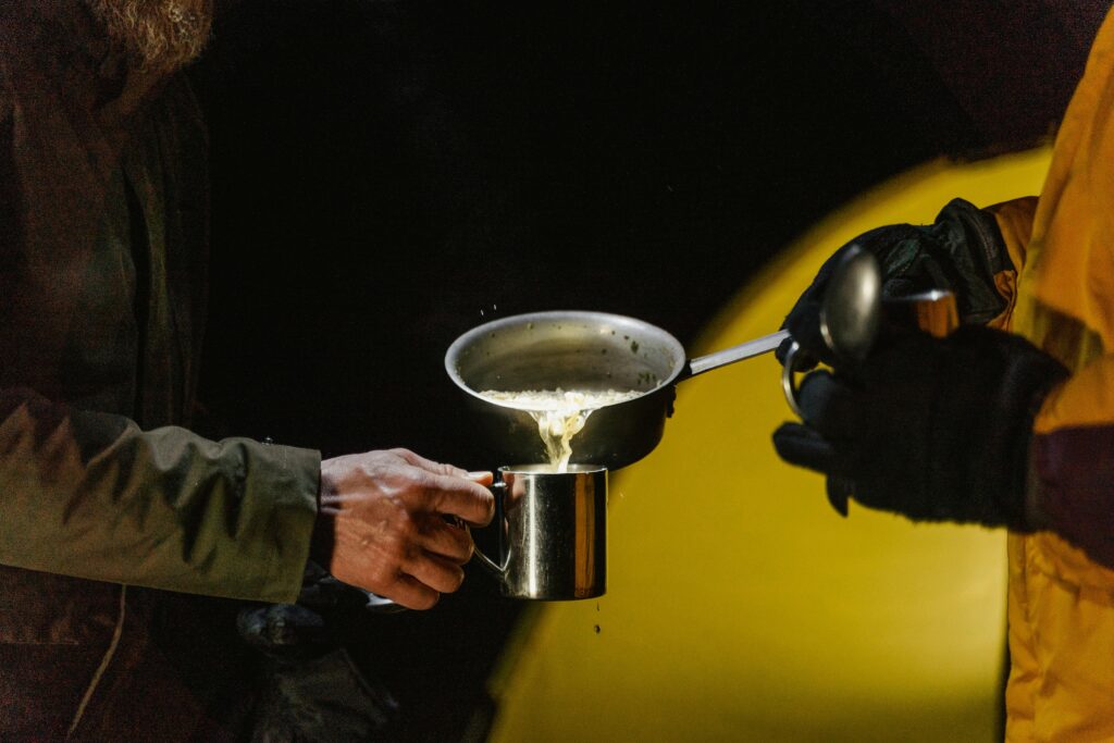 Two hands pour steaming liquid from a pan into a metal mug, surrounded by darkness and a softly lit yellow tent.