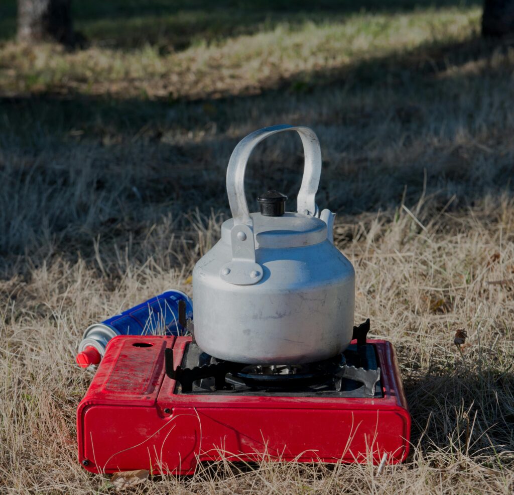 A silver kettle on a red portable stove, set on dry grass in a wooded area, with a small blue canister nearby.