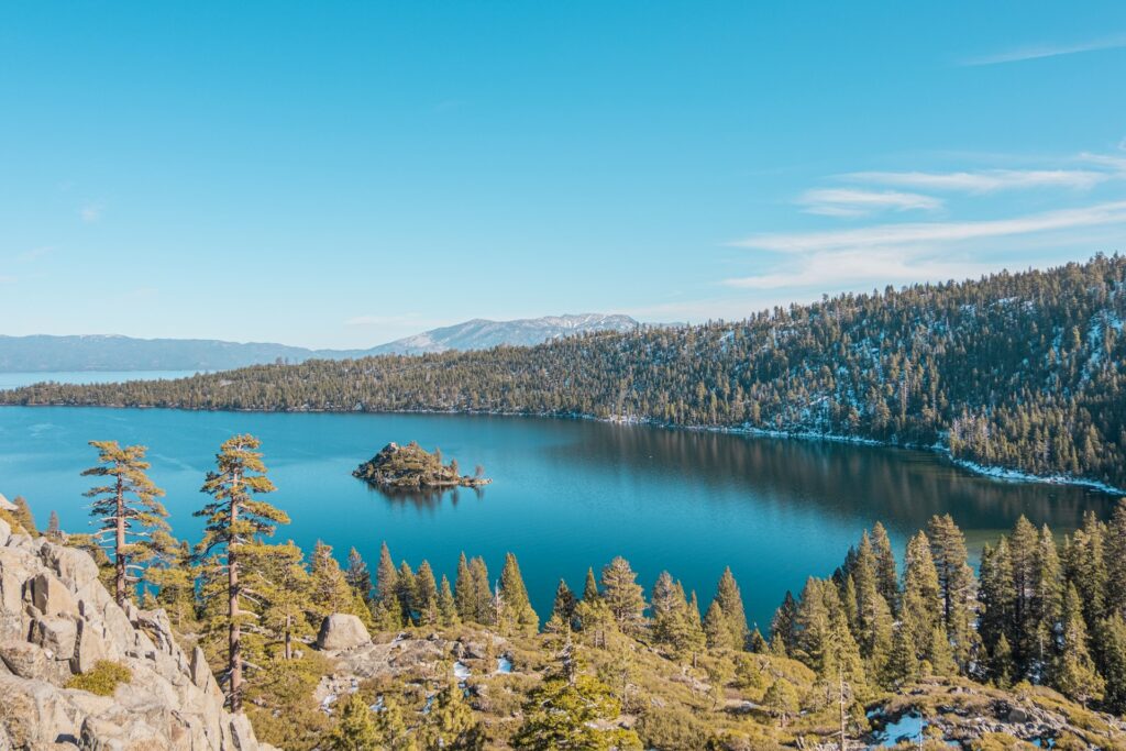 A tranquil lake scene with a small island, surrounded by lush forests and distant mountains under a clear blue sky.